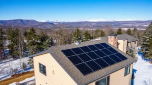 Aerial view of modern residential rooftop with solar panels installed, winter New Hampshire landscape with snow-covered trees and mountains in background, clear blue sky