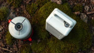 Overhead view of white gas metal fuel container sitting on forest moss with camping stove nearby, natural daylight, photorealistic, no text or labels visible