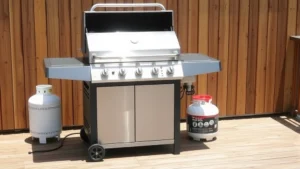Modern stainless steel gas grill on wooden deck with propane tank, bright sunlight, natural wood fence background, no text or labels visible