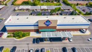 Aerial view of a modern Sam's Club warehouse with a busy fuel station in the foreground, multiple cars refueling under covered pumps, clean parking lot with marked spaces, bright daylight, suburban setting