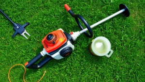 Overhead view of a gas-powered weed eater on green grass with trimmer line visible, surrounded by maintenance tools and fresh gasoline in a metal container, natural daylight